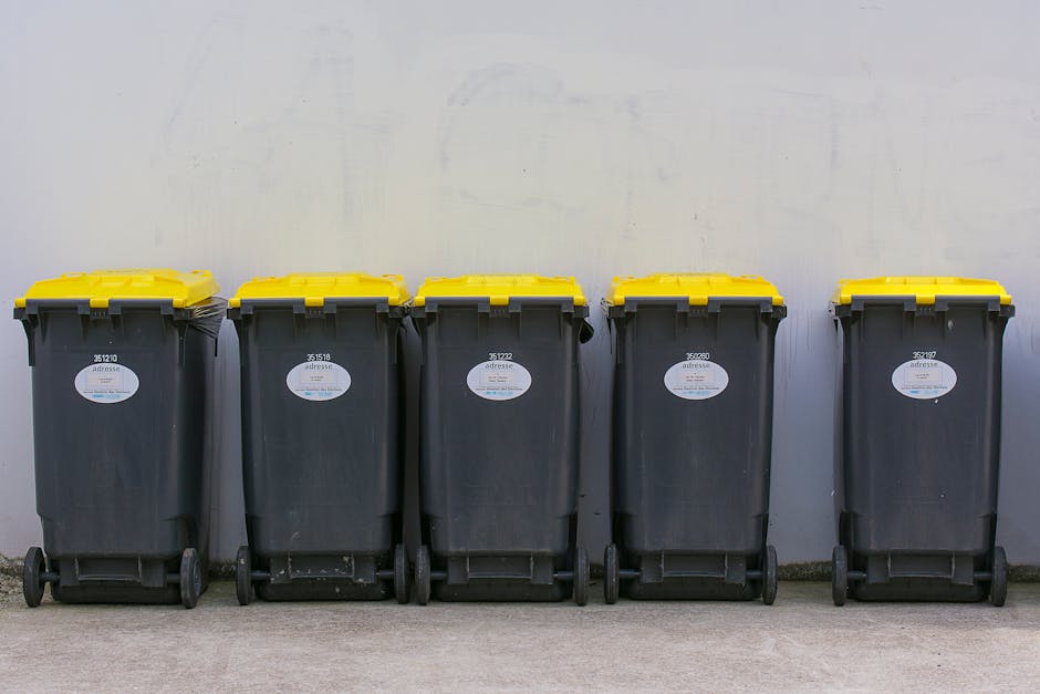 A worker dressed in a high-visibility yellow and red uniform operates a red commercial rubbish collection vehicle positioned on the side of a street. The back of the vehicle is open, revealing mechani